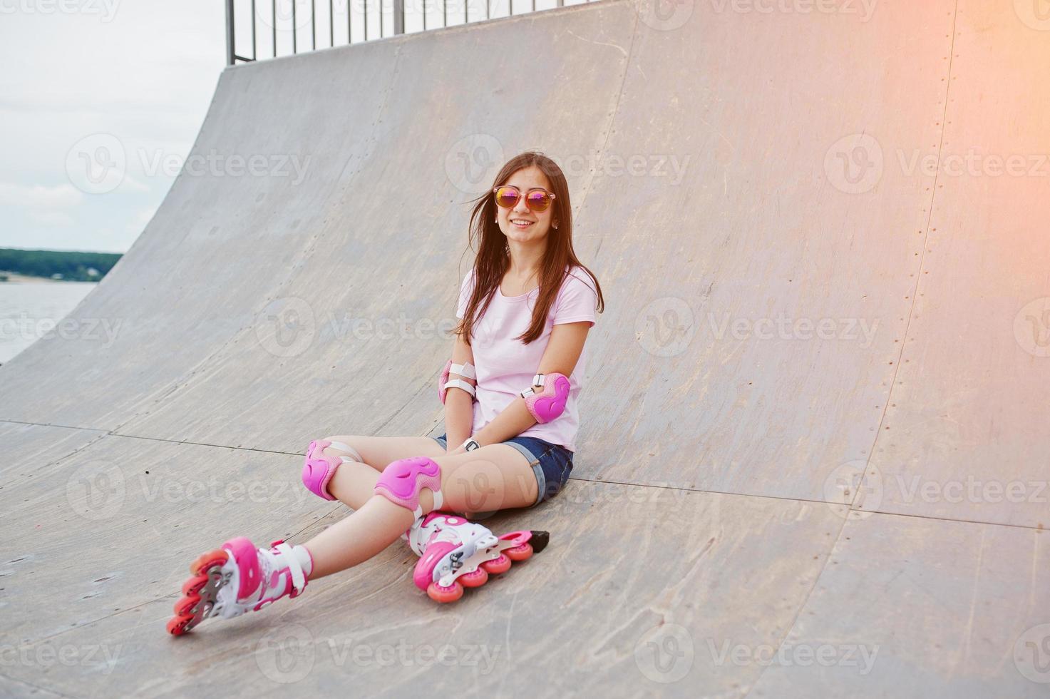 retrato de una hermosa chica sentada en la pista de patinaje al aire libre en pantalones cortos, camiseta, gafas de sol y patines. foto