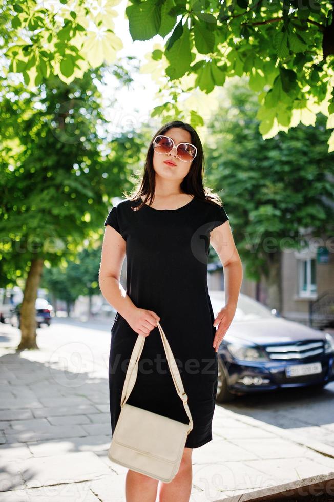 Brunette business girl at black dress on sunglasses with handbag at hand posing at street of city, background car on road. photo