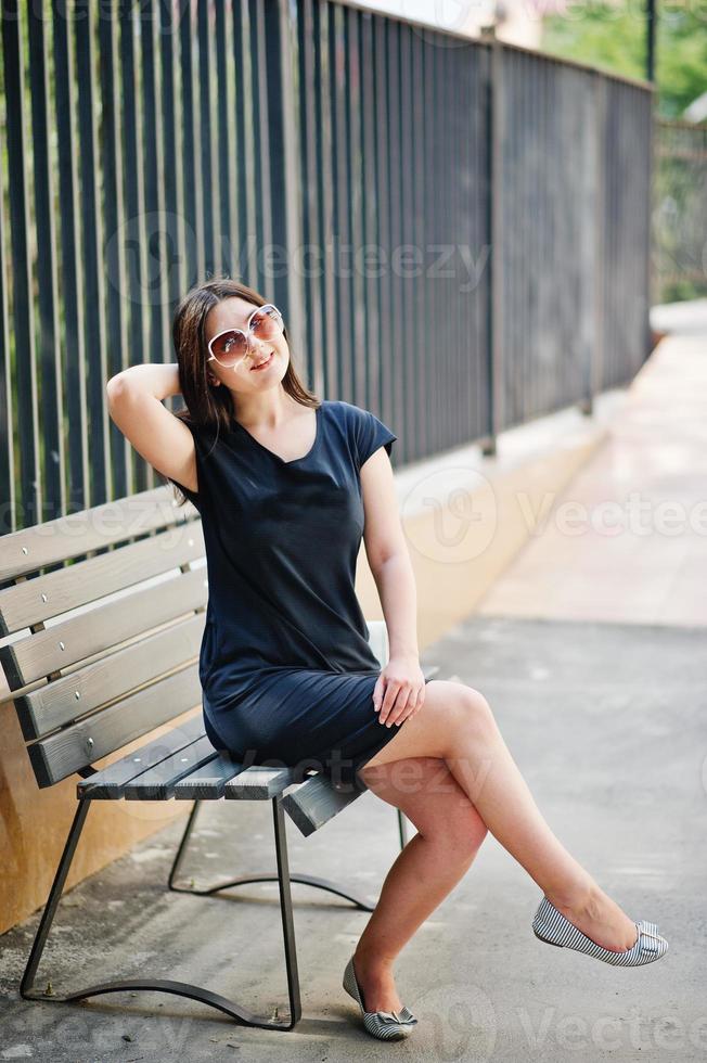 Brunette girl at black dress on sunglasses sitting on bench and posing at street of city. photo