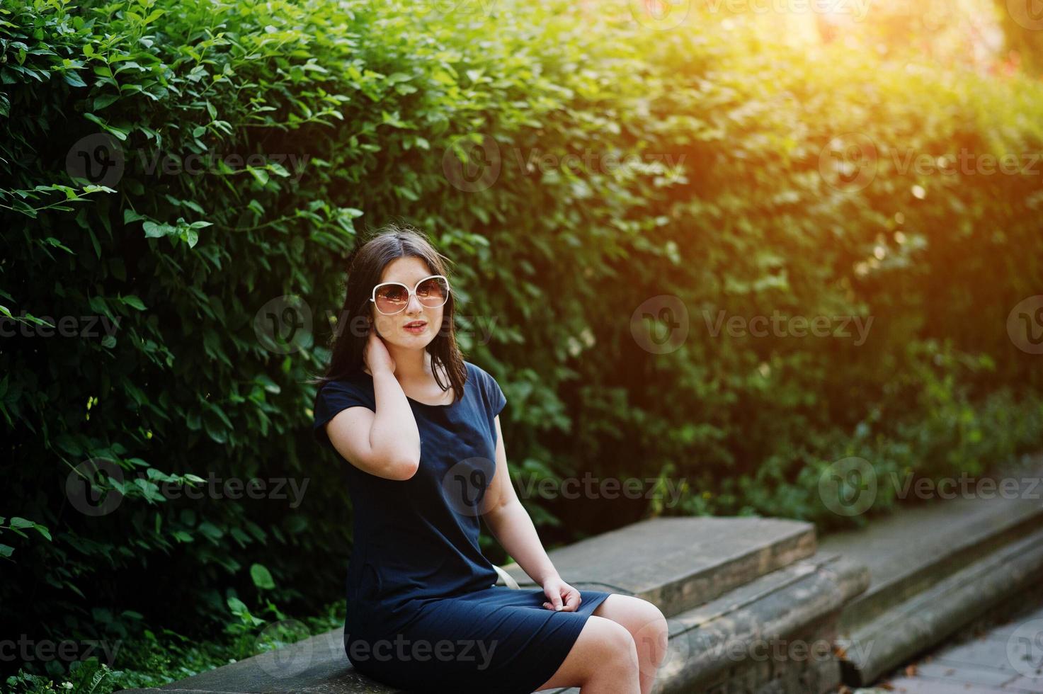 Brunette business girl at black dress on sunglasses sitting at border against bushes, posing at street of city. photo