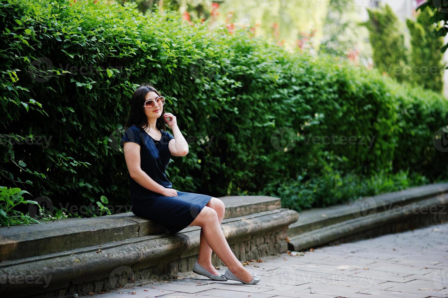 Brunette business girl at black dress on sunglasses sitting at border against bushes, posing at street of city. photo