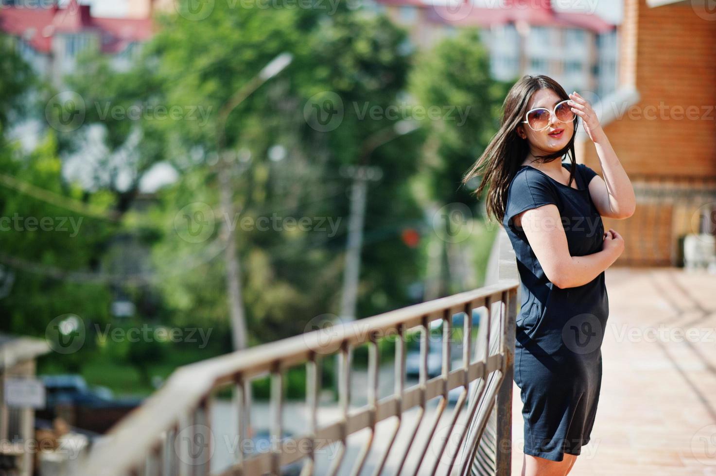 chica morena con vestido negro, gafas de sol posando en la calle de la ciudad. foto