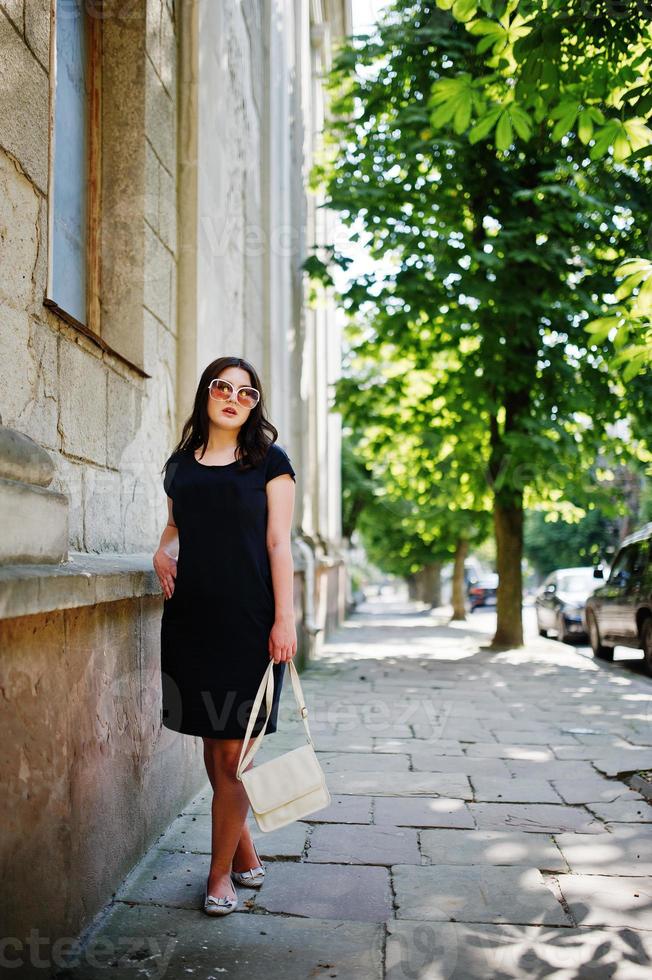 Brunette girl at black dress on sunglasses with handbag at hand posing at street of city. photo