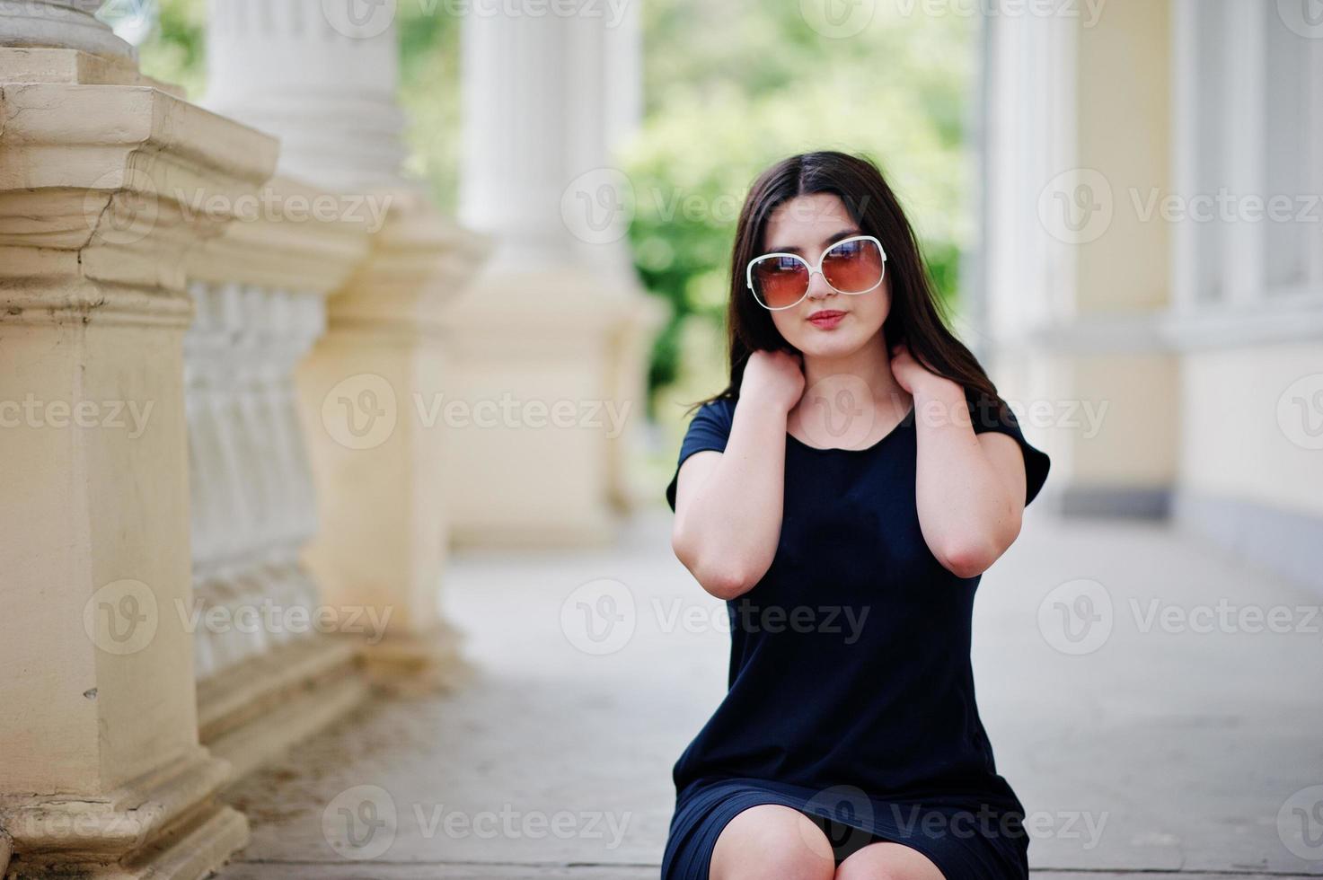 Brunette girl at black dress, sunglasses sitting on stairs of old vintage house, posing at street of city. photo