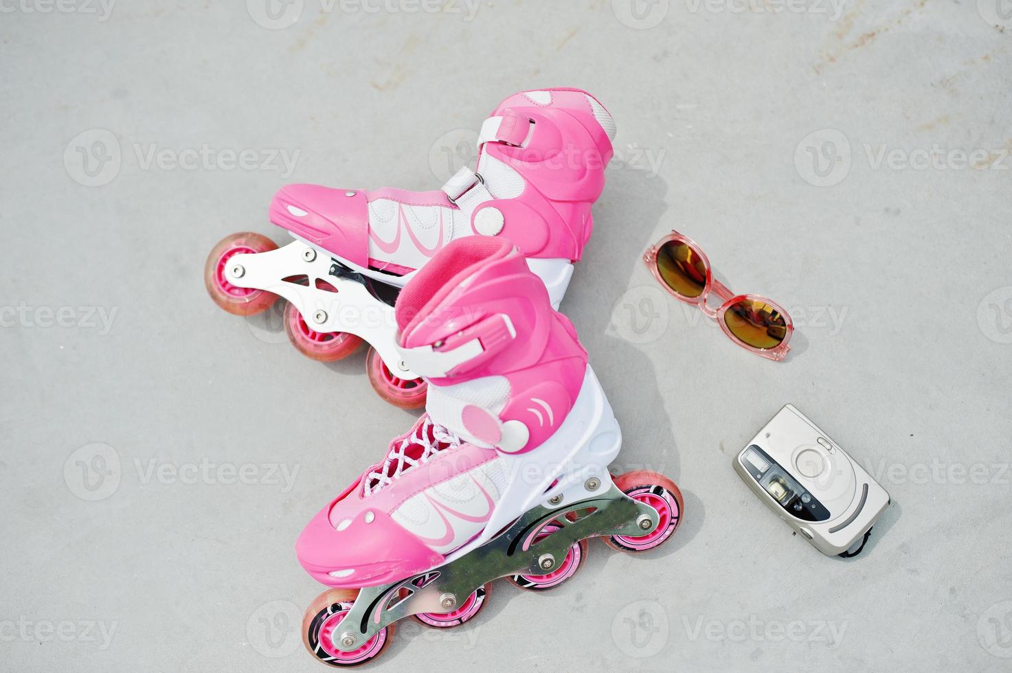 Close-up photo of pink rollerblades laying along with an antique camera, sunglasses and a pink cap.