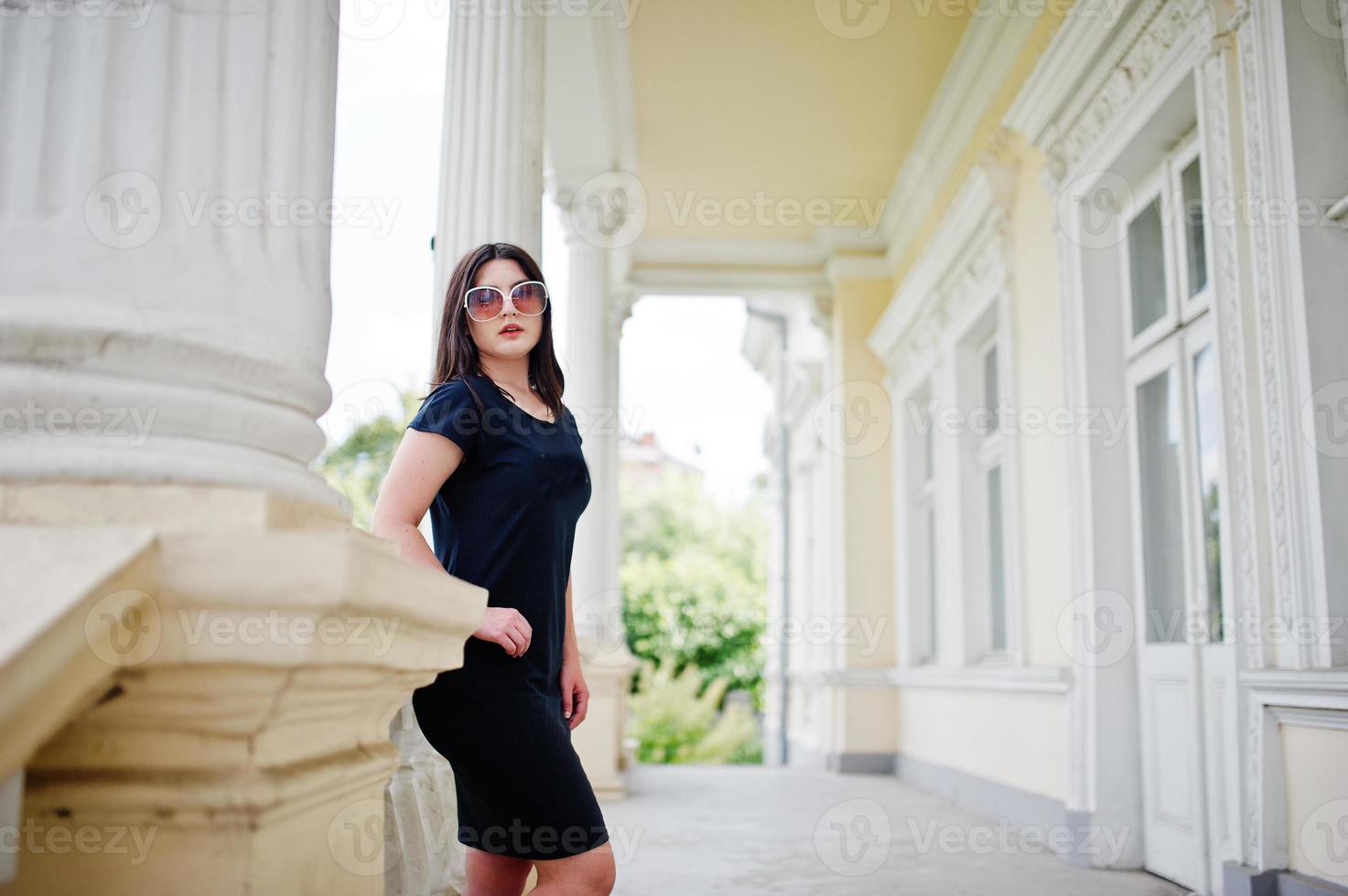 Brunette girl at black dress, sunglasses posed against old vintage house, at street of city. photo