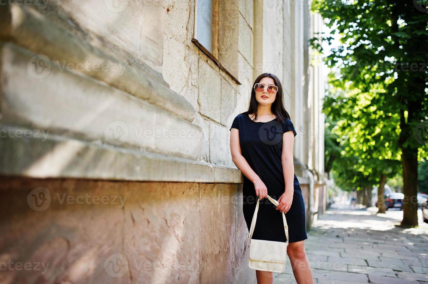 Brunette girl at black dress on sunglasses with handbag at hand posing at street of city. photo