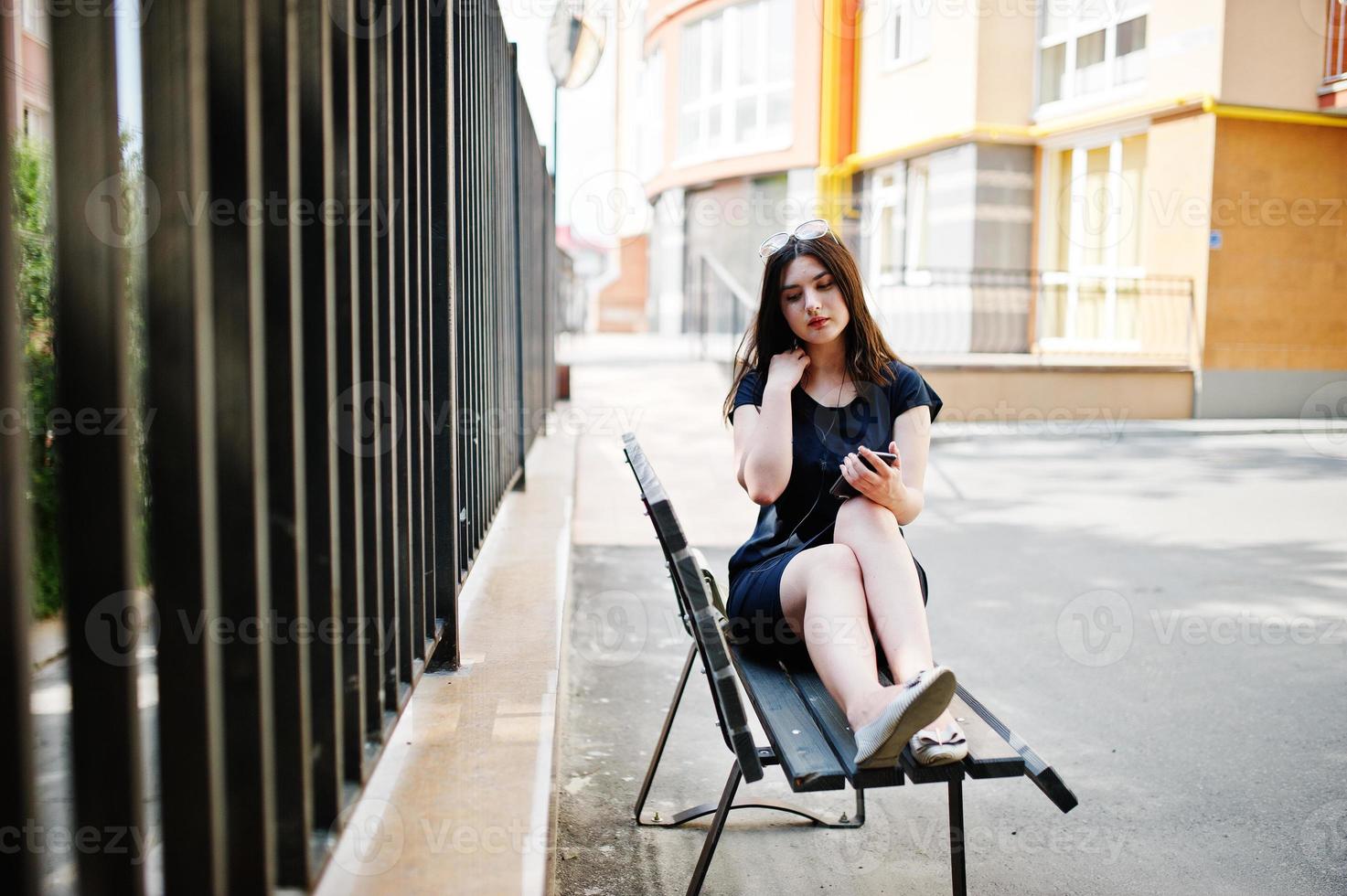 chica morena con vestido negro, gafas de sol sentada en un banco, escuchando música desde el teléfono con auriculares y posando en la calle de la ciudad. foto