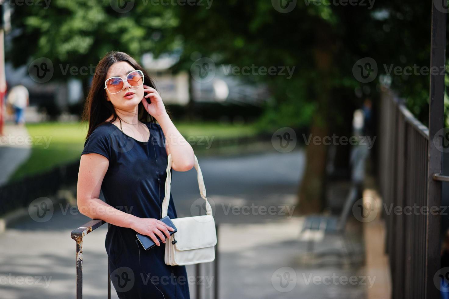 chica morena con vestido negro, gafas de sol escuchando música desde el teléfono con auriculares y posando en la calle de la ciudad. foto