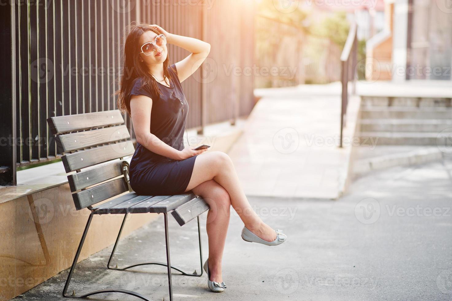 Brunette girl at black dress, sunglasses sitting on bench, listening music from headphones phone, and posing at street of city. photo