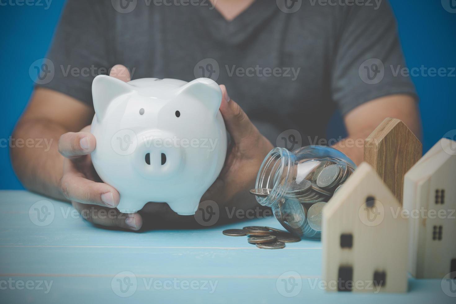 A man holding a white pig piggy bank in hand with model home wood, and coins in the glass. house trading concept. Housing, bank loans to buy a house, real estate investment, real estate business. photo