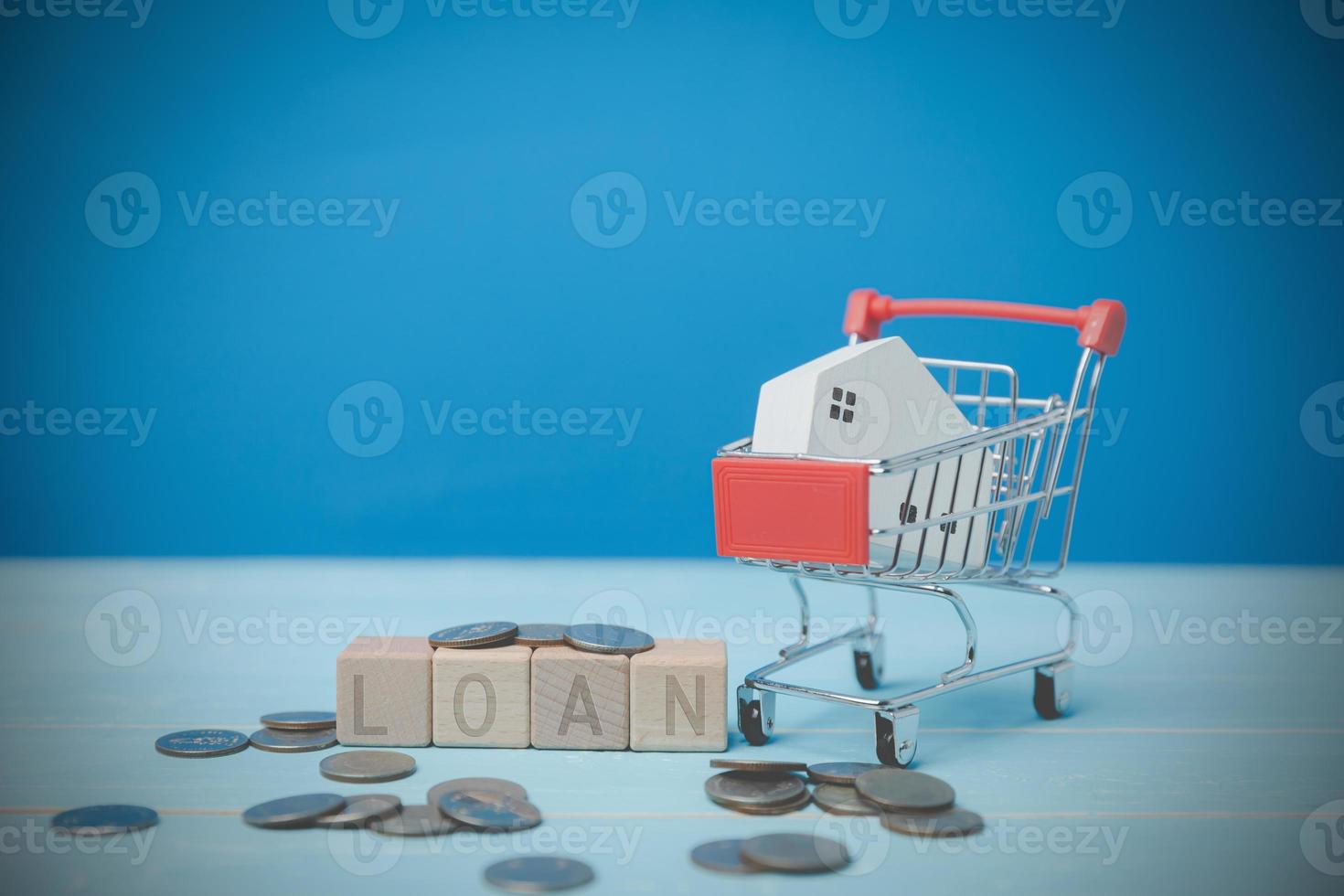 A mini wood house model is in a shopping cart with text LOAN on blocks wooden on a blue desk. Real estate trading ideas, savings for the future, banking, finance, and accounting. photo