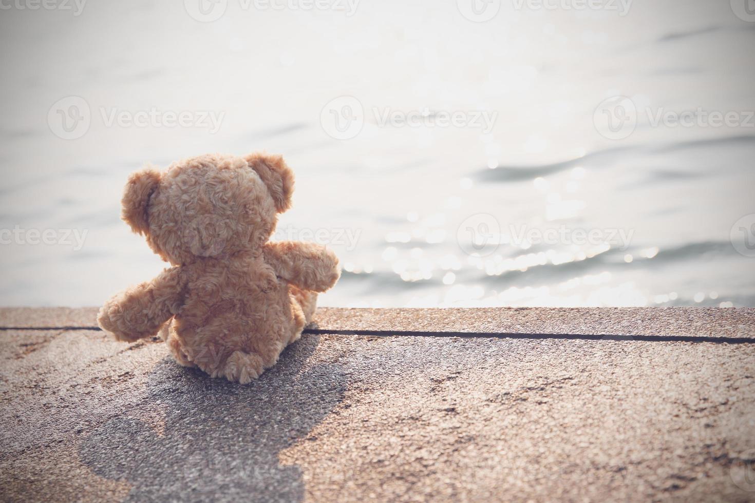 A sad teddy bear sits on a bridge alone looking at the sea in lonely with copy space. Toy, doll, for child. photo