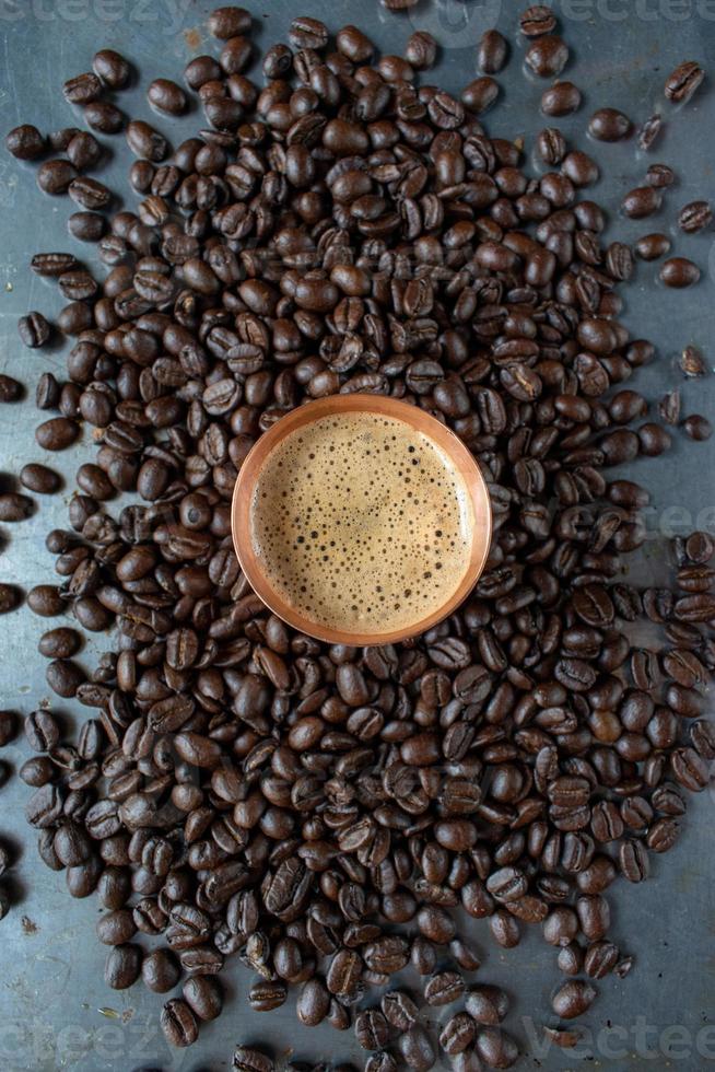 taza de cobre llena de café espresso en el centro de granos de café crudos esparcidos sobre una mesa rústica plana foto