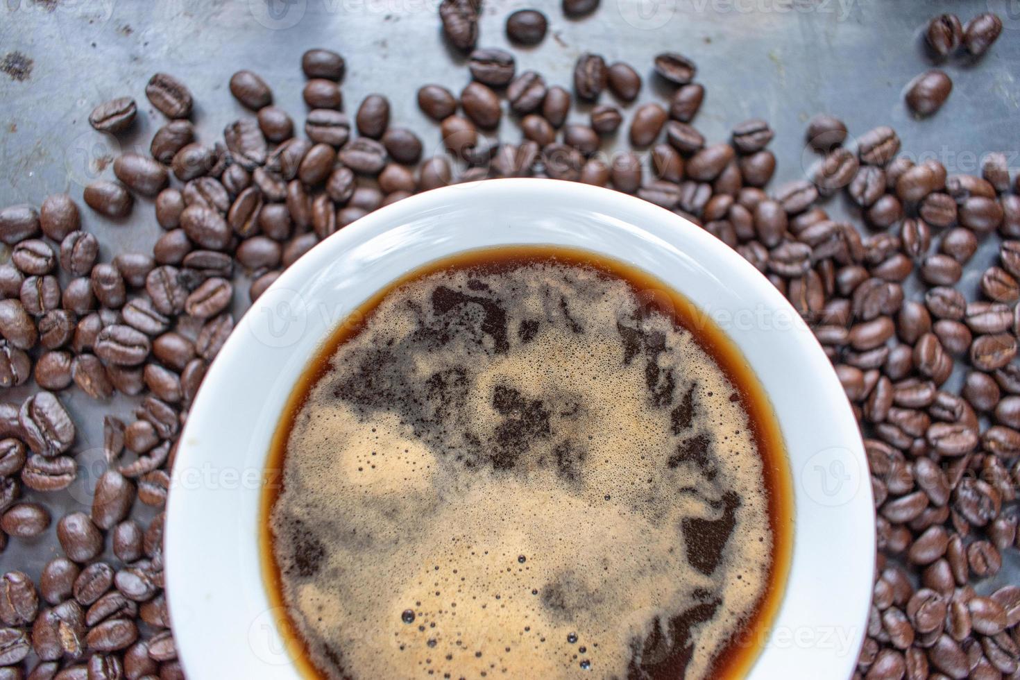 taza de café blanca llena de café fuerte en el centro de granos de café crudos esparcidos sobre una mesa rústica plana foto