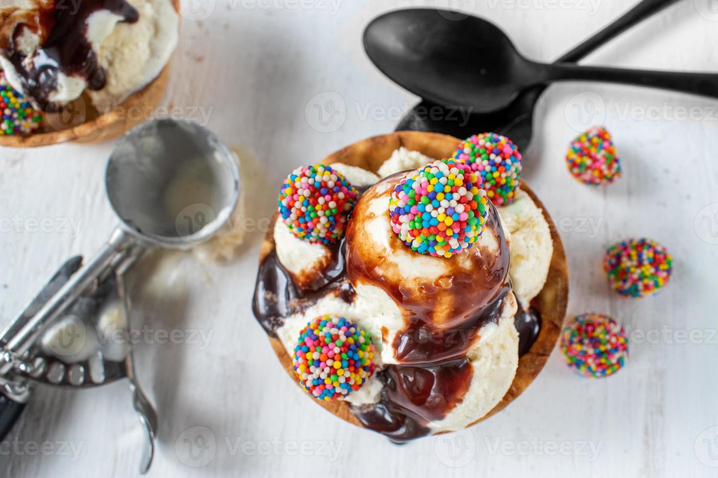 vanilla ice cream scoops with colorful candy pieces and chocolate syrup in waffle cone bowl on rustic white background photo