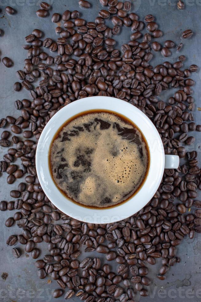 taza de café blanca llena de café fuerte en el centro de granos de café crudos esparcidos sobre una mesa rústica plana foto