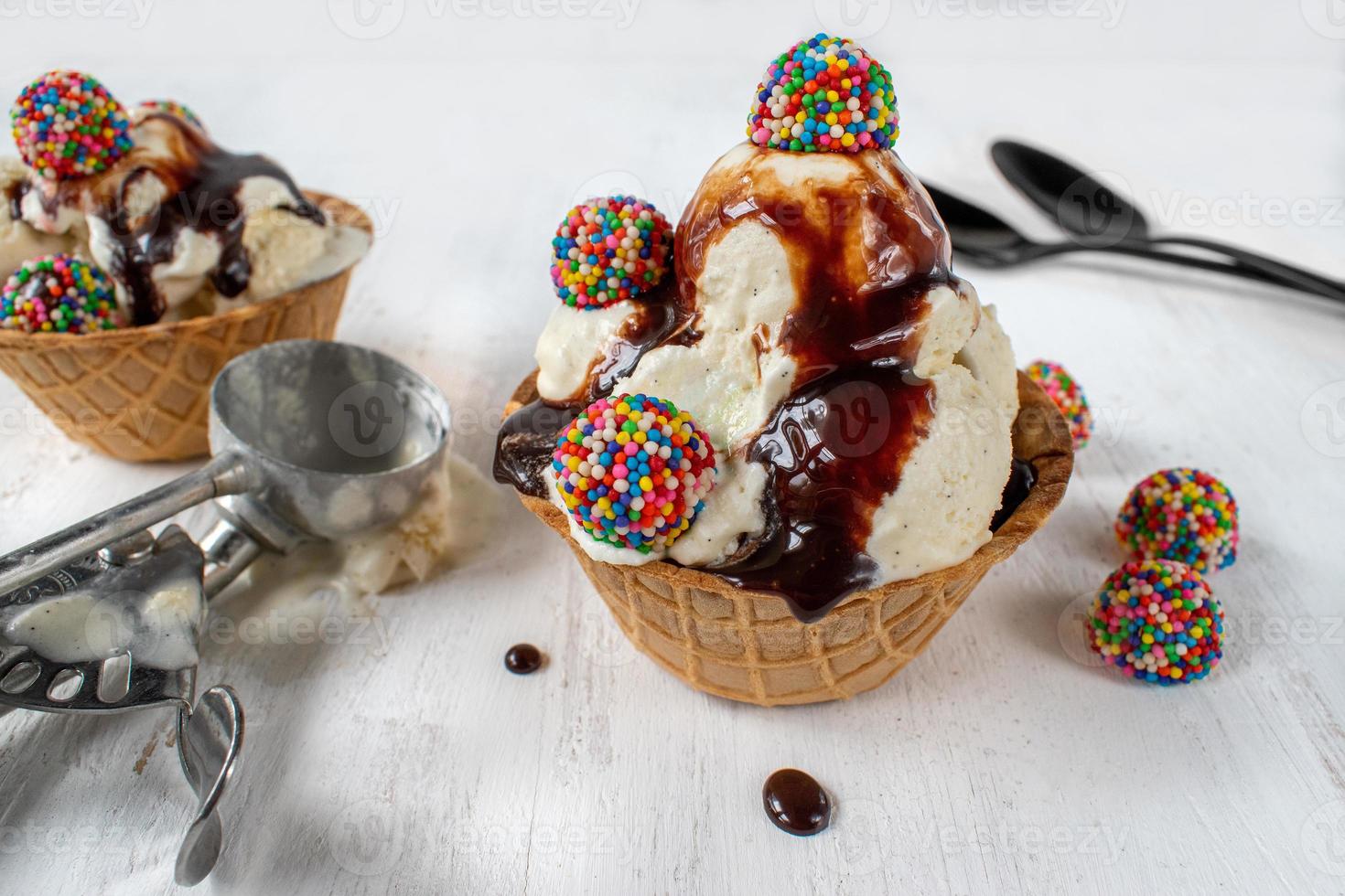 bolas de helado de vainilla con piezas de caramelos de colores y jarabe de chocolate en un tazón de cono de gofre sobre fondo blanco rústico foto