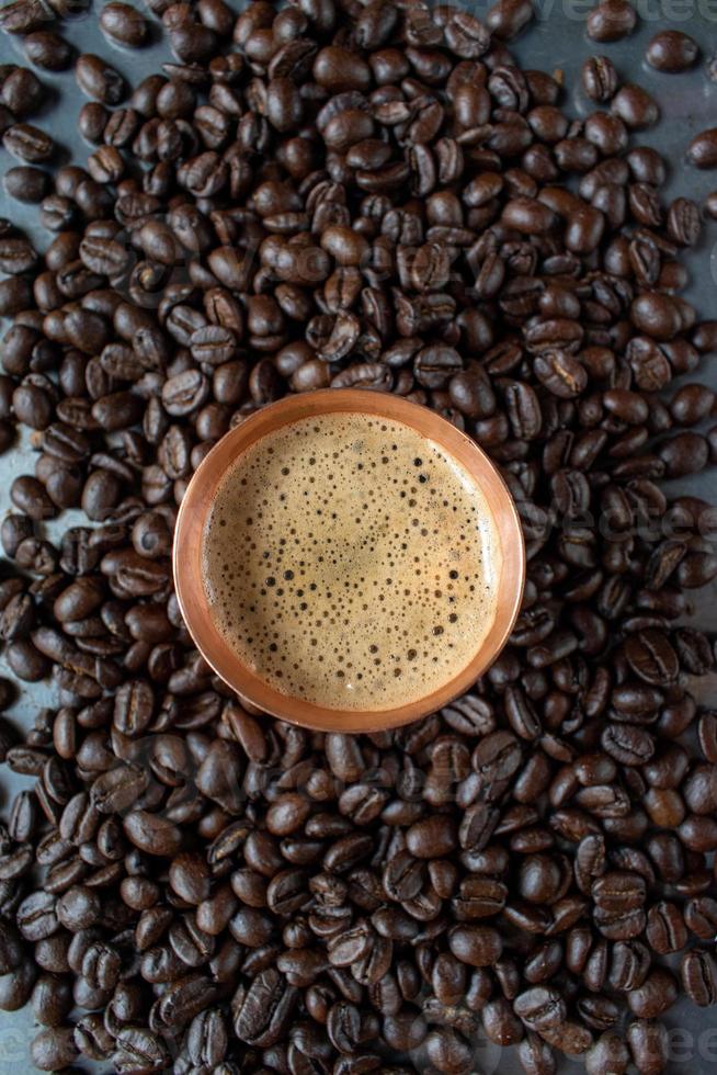 taza de cobre llena de café espresso en el centro de granos de café crudos esparcidos sobre una mesa rústica plana foto
