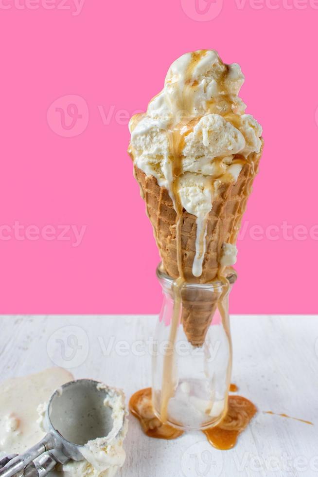 bolas de helado de vainilla derritiéndose con salsa de caramelo goteando sobre un cono de gofre sobre un divertido fondo rosa vibrante foto