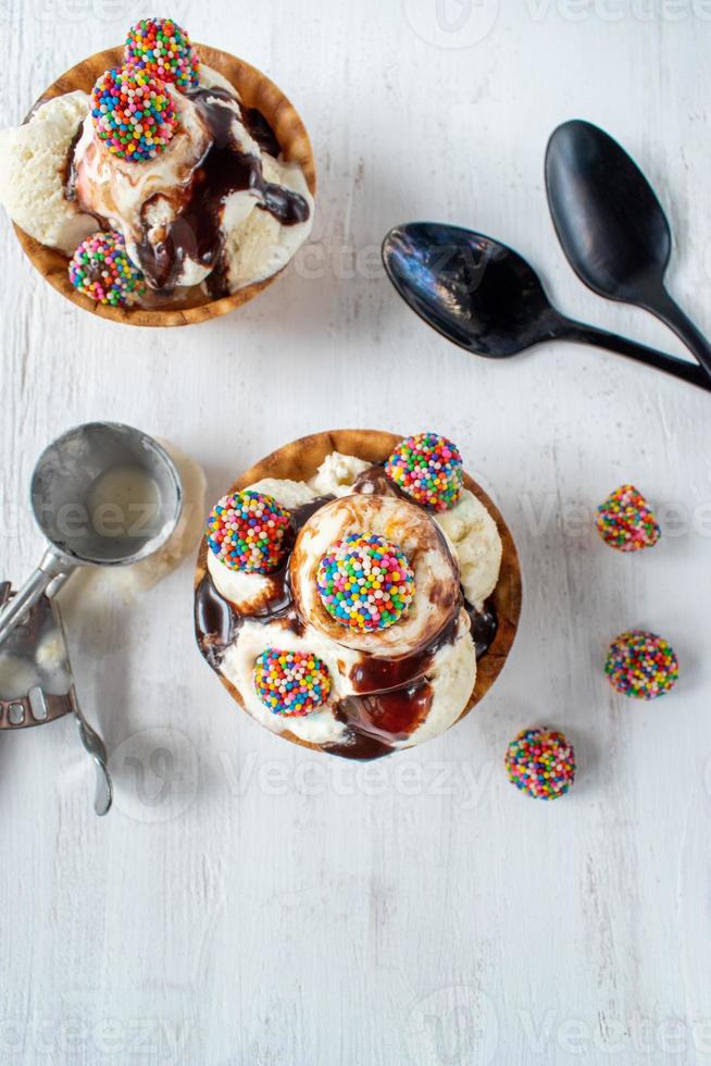 vanilla ice cream scoops with colorful candy pieces and chocolate syrup in waffle cone bowl on rustic white background photo