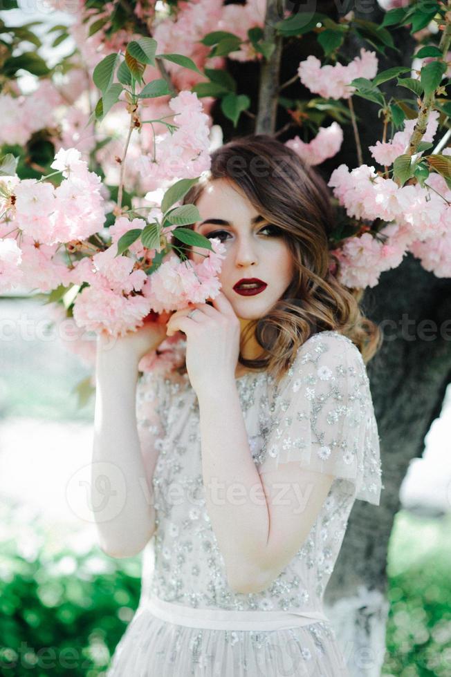 muy hermosa chica la novia bajo un velo, vestido de novia beige cerca del árbol sakura está mirando hacia abajo, naturaleza foto