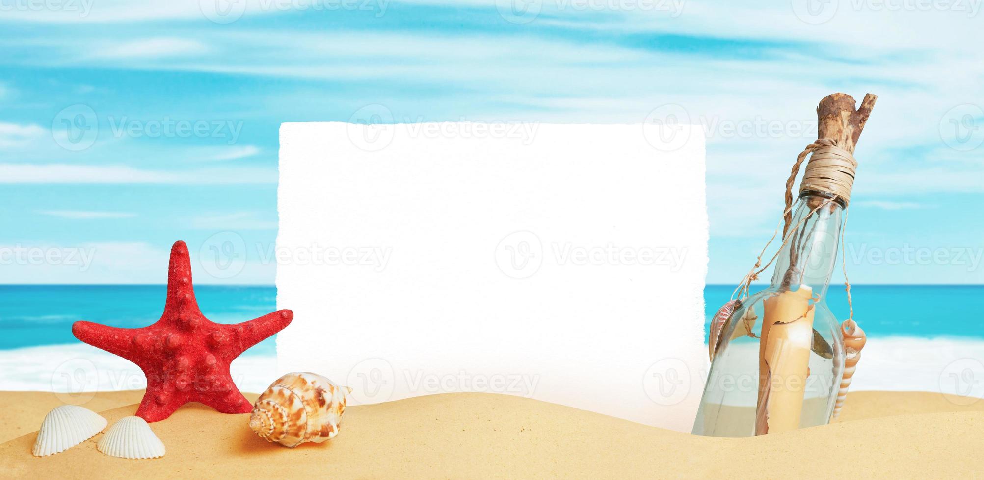 White paper stuck in the sand of the beach, surrounded by starfish, shells and a bottle with a message. Clean surface for promo text photo