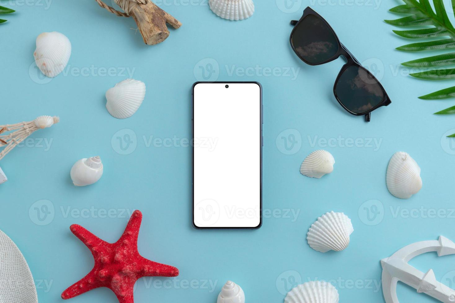 Phone mockup surrounded by shells, starfish, sunglasses, palm leaves on blue surface. Isolated screen for app design presentation. Summer travel composition. Top view, flat lay photo