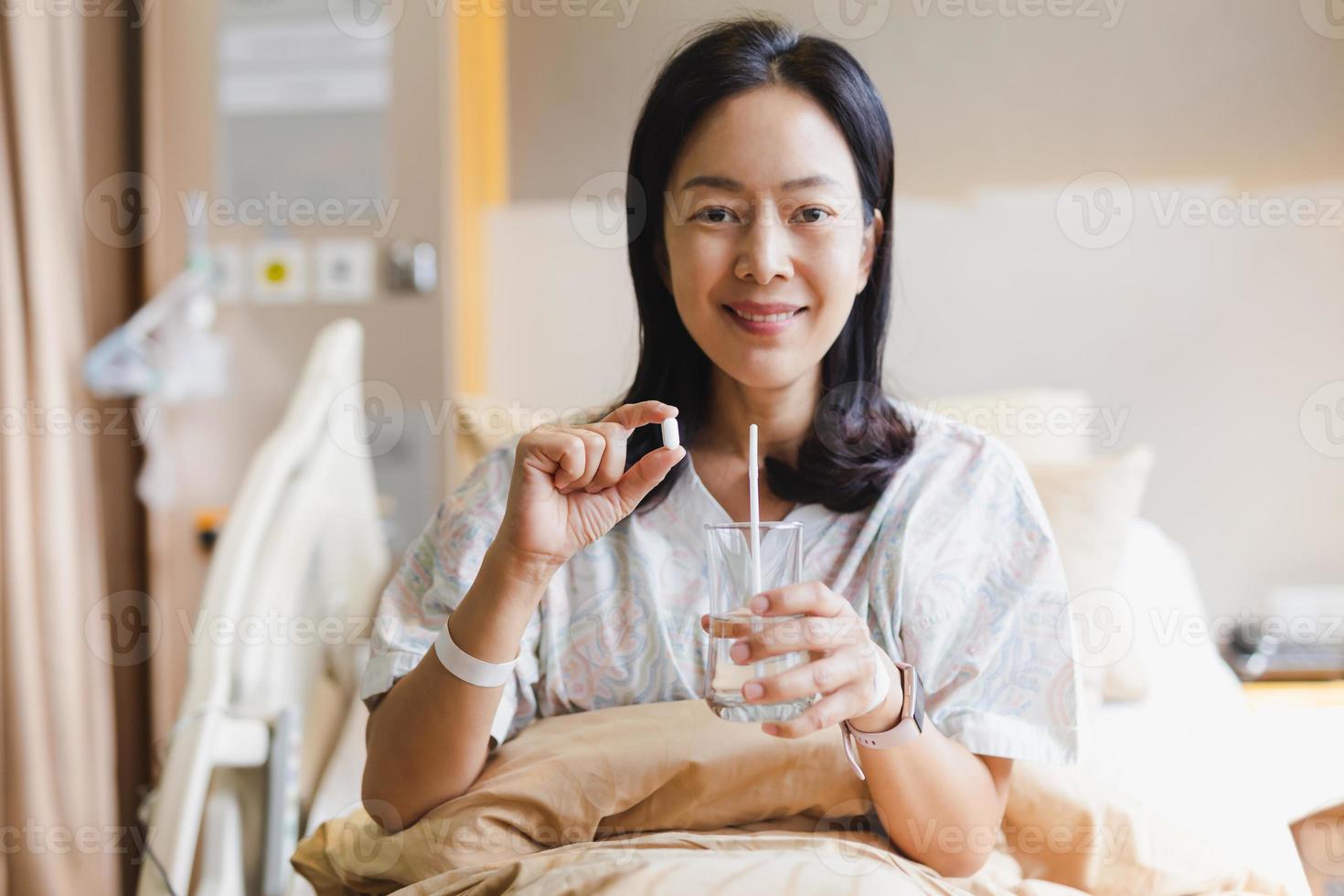 Woman holding white pills and a glass of water while sitting on a bed in hospital. photo