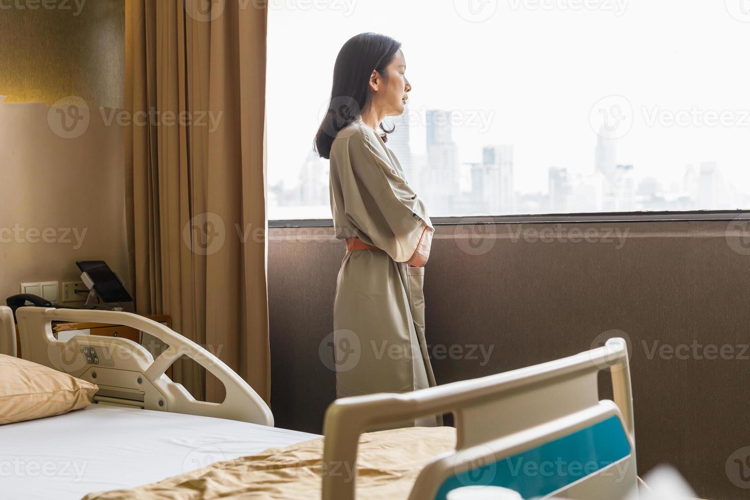 Una paciente asiática deprimida de pie está mirando por la ventana de la sala del hospital. foto