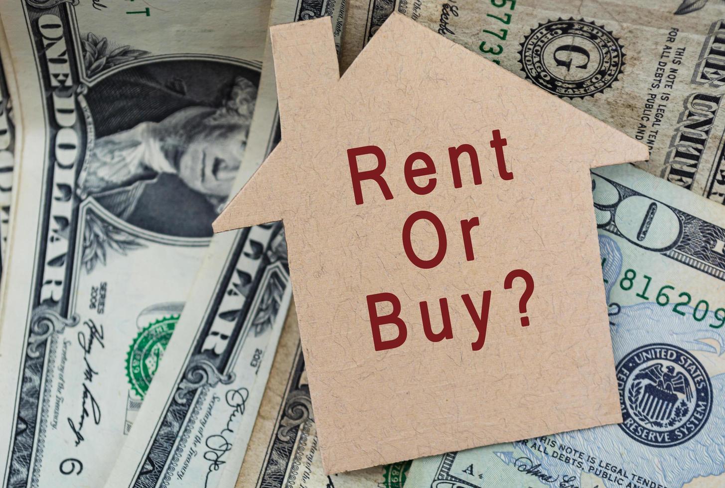 Rent or buy with question mark text on on brown paper house model with dollar banknotes on dark wooden table photo