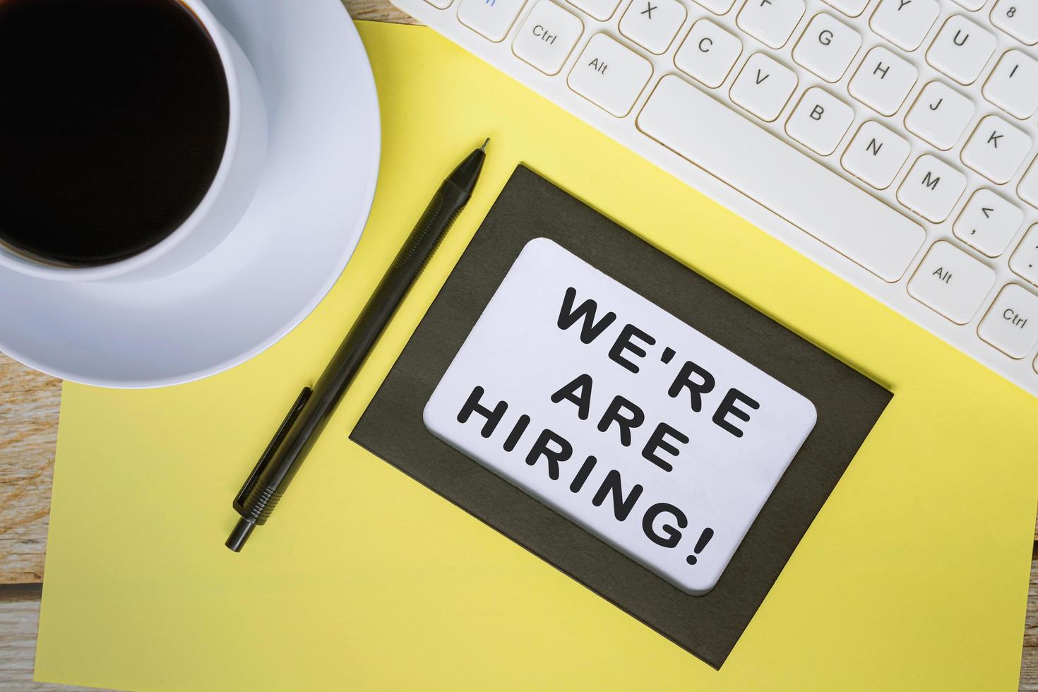 We're are hiring text on white note with yellow sheet on wooden desk. photo