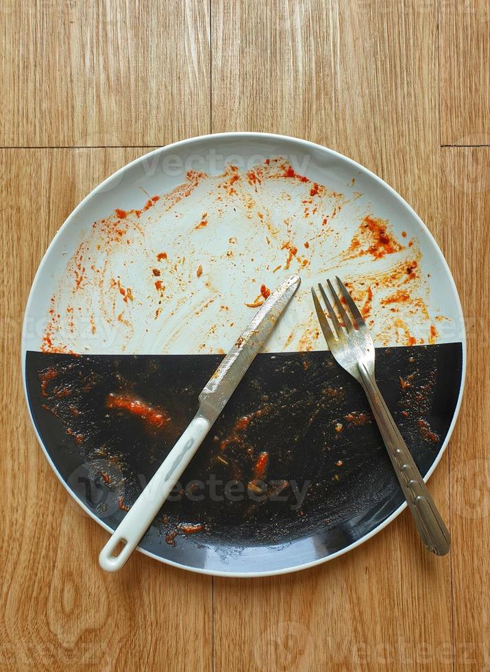 Top view of dirty black and white dish, fork, and knife on wooden table. Leave over sauce smeared on a plate. For use in soap, detergent, liquid, sponge, cloth, washing machine advertising. photo