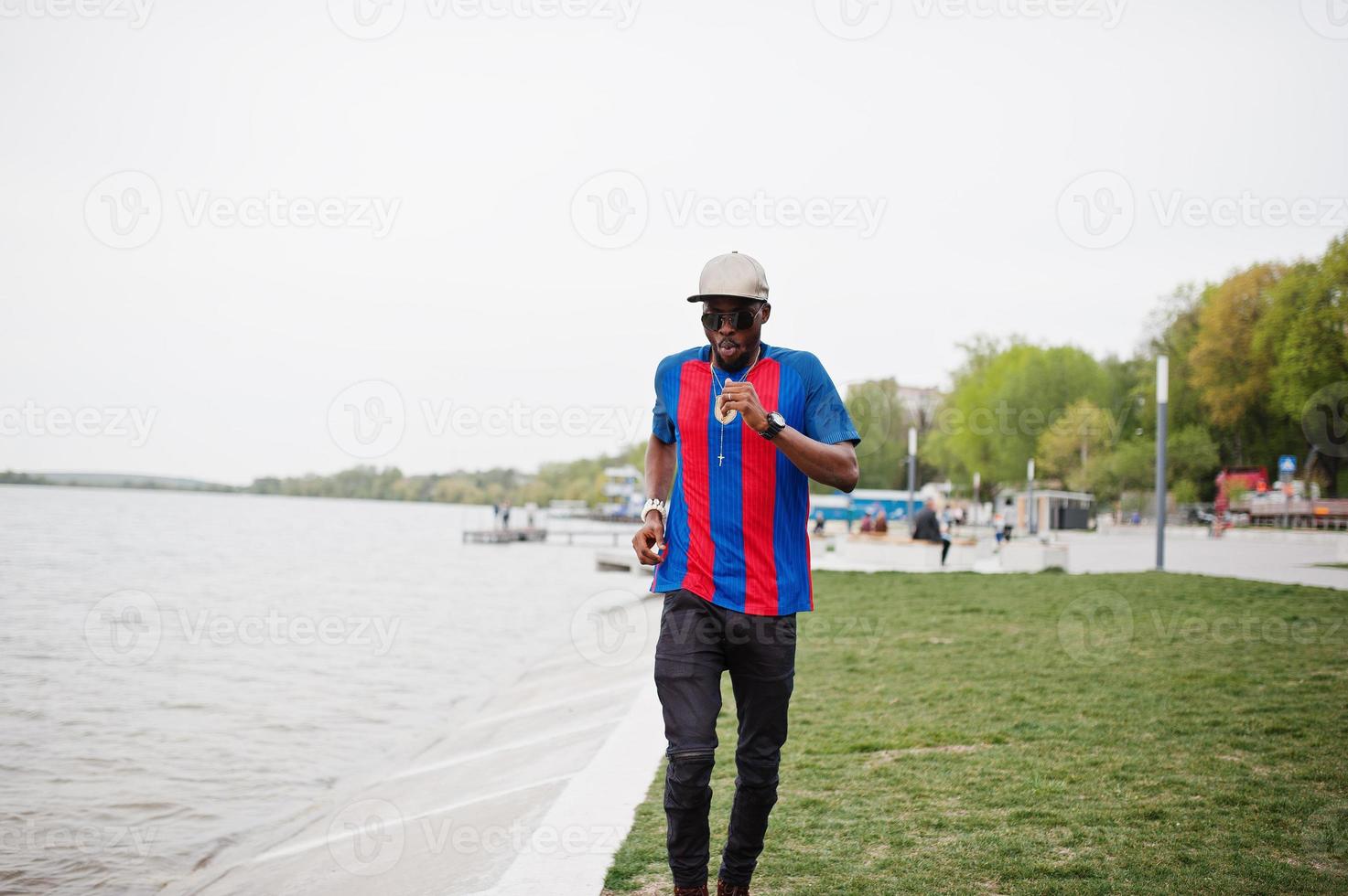 Stylish african american boy running against lake wear at cap, football t-shirt and sunglasses. Black sports man portrait. photo