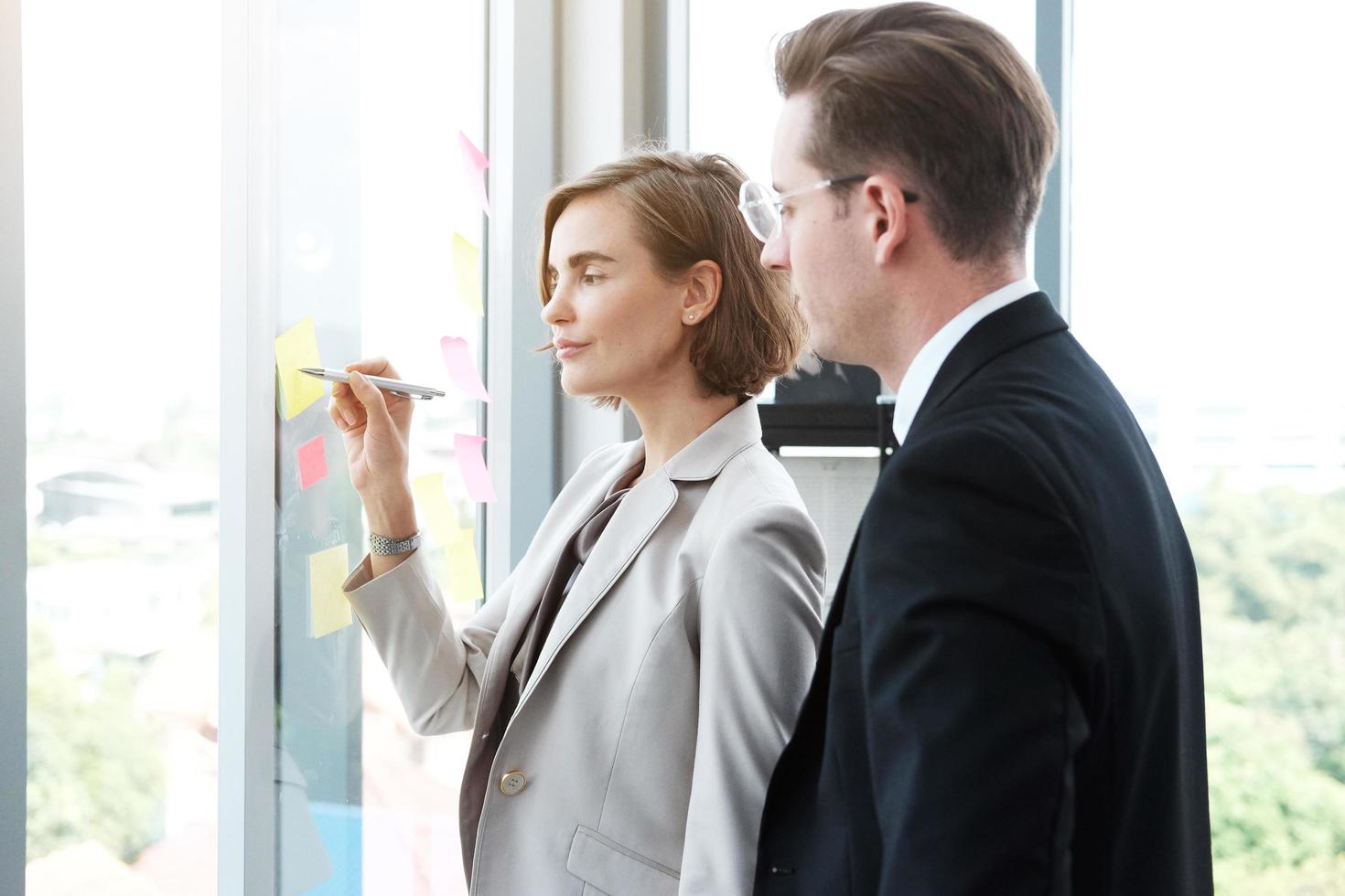 Successful businessman and businesswoman writing good ideas on sticky notes pasted on glass wall  in modern office meeting. photo