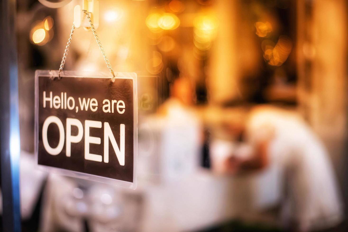 la señalización de la tienda está abierta del restaurante colgada en la puerta y la gente de fondo borrosa horas felices después del cierre de la epidemia de covid-19, texto en un moderno letrero negro de plástico hola, estamos abiertos en el café. foto