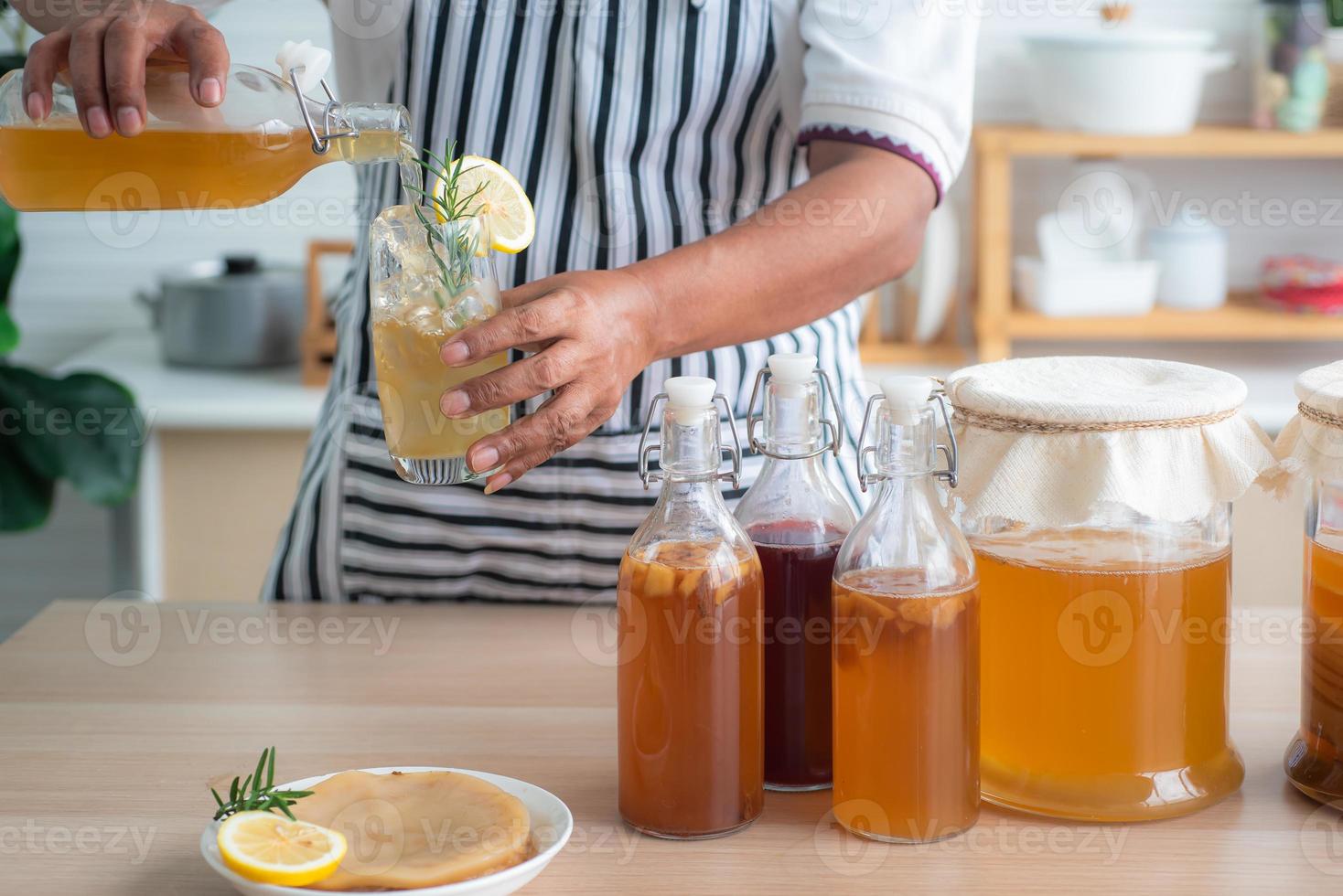 Chef's hand holds a bottle and a glass of Homemade fermented kombucha tea, variety of flavors in bottles and glass jars arranged mix with fruit, scoby on wooden table. Healthy natural probiotic drink. photo