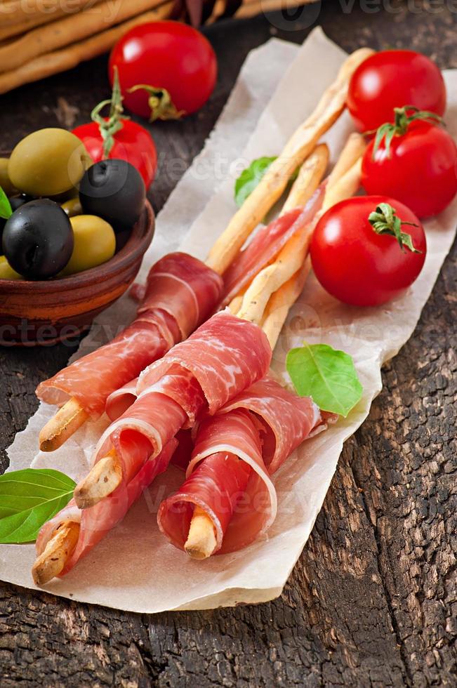 Grissini bread sticks with ham, olives, basil on old wooden background photo