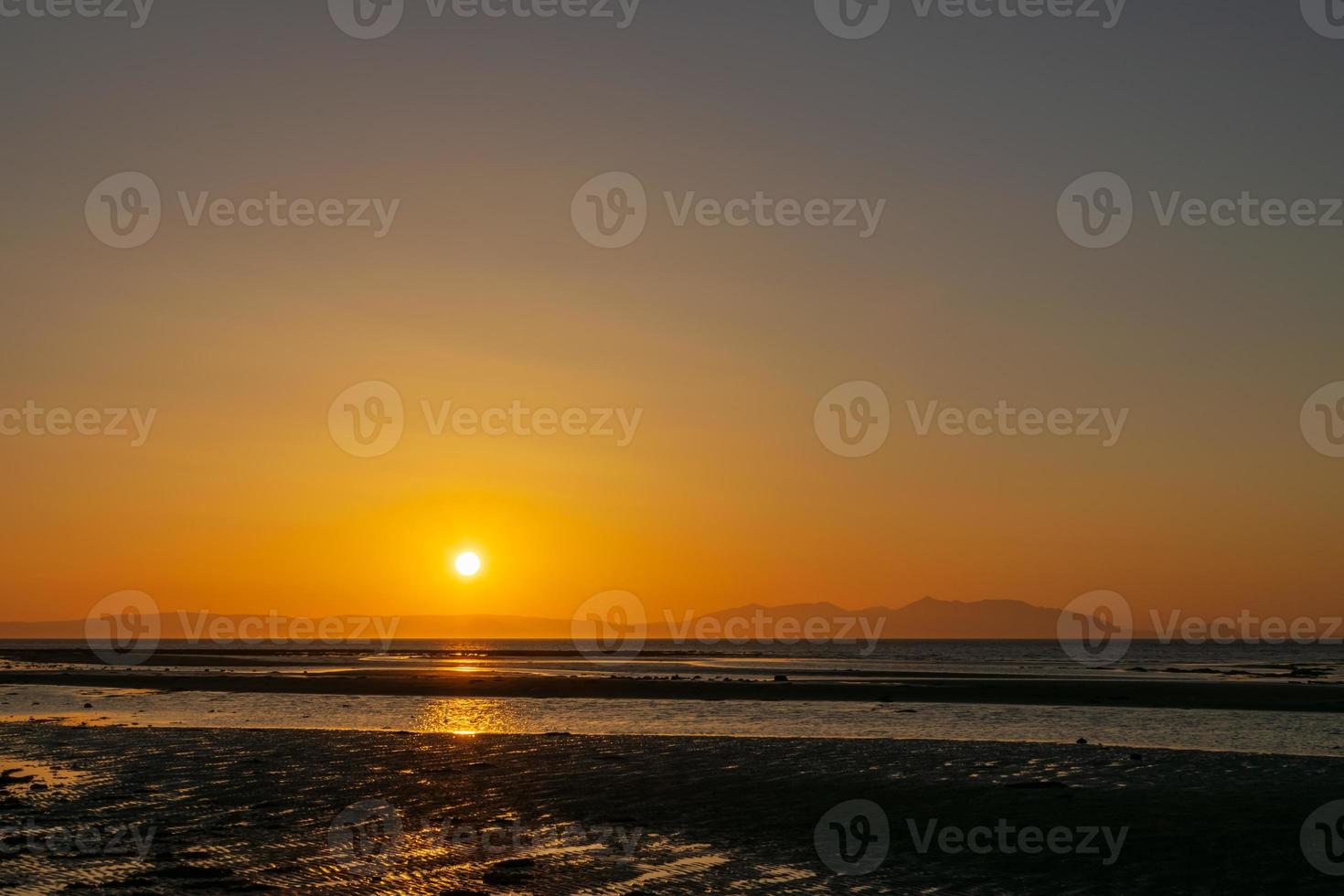 puesta de sol sobre la playa y la montaña, línea del horizonte reflejo de sombreado amarillo y naranja en el agua y la arena. enfocándose en la sombra de la superficie. paisaje naturaleza paisaje fondo. foto