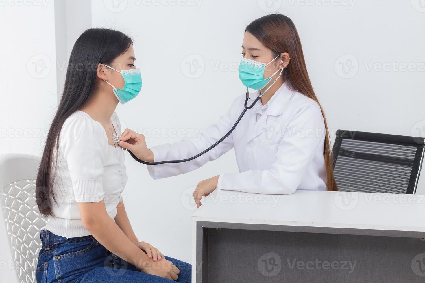 Asian female doctor examines symptom of her patient while both wear medical face mask to prevent infection disease COVID 19 in hospital. photo