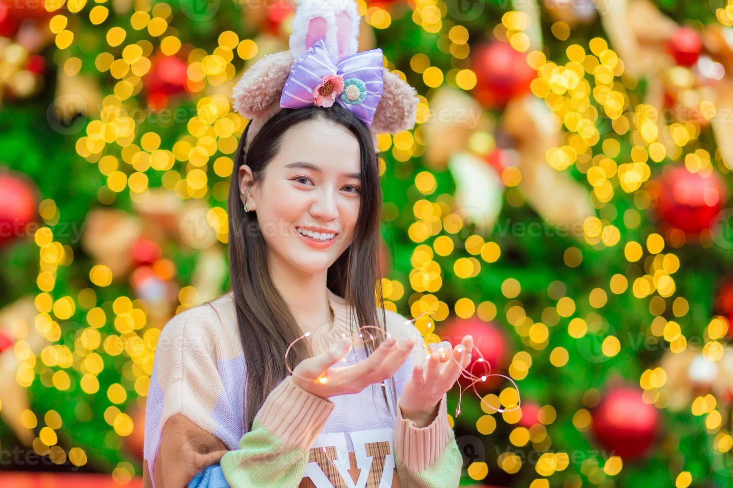 una mujer asiática con un suéter colorido se para frente al árbol de navidad. con bokeh como fondo en el tema de celebrar el año nuevo foto