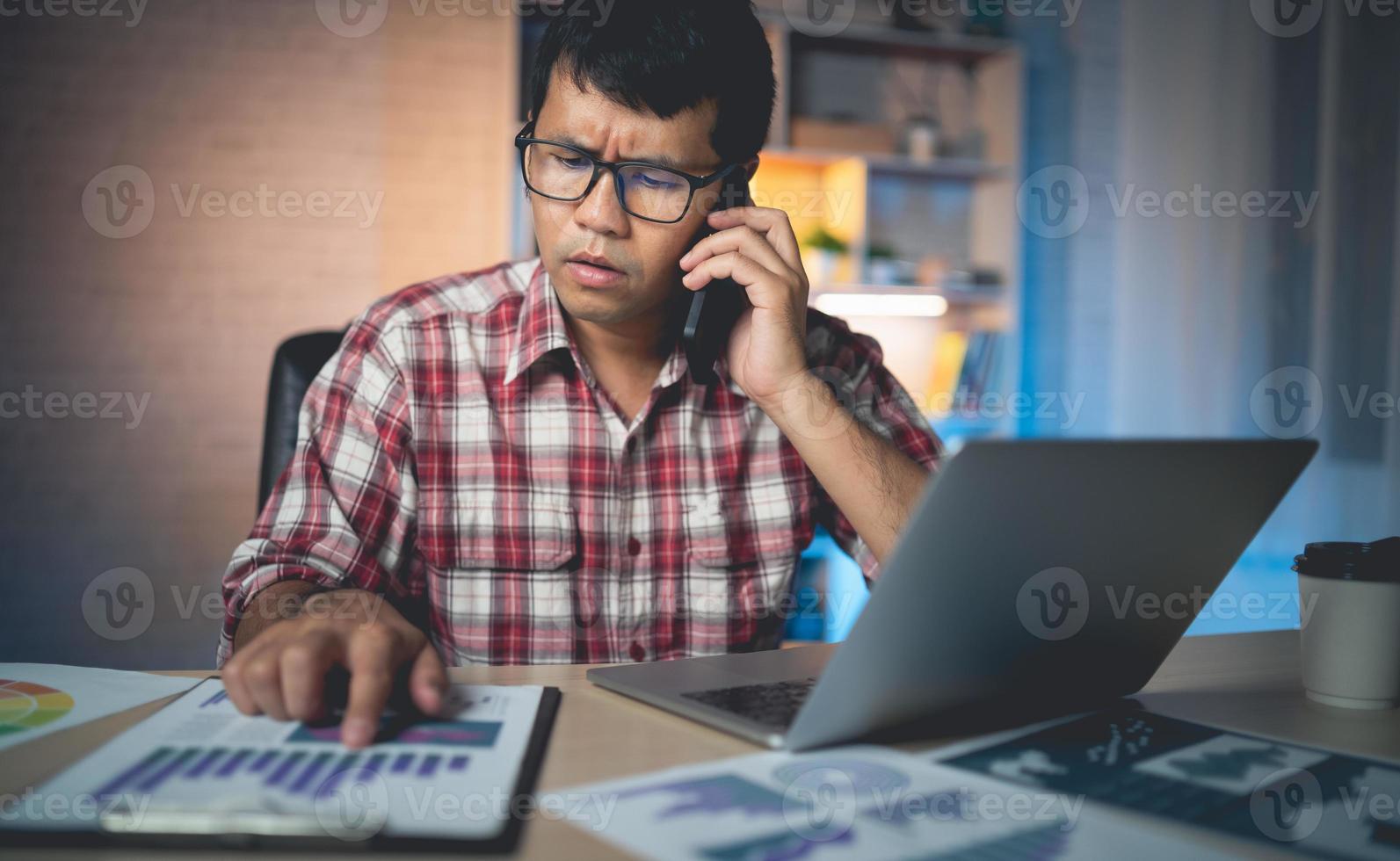 Freelance asian man with stressed and worried expression Talking on mobile phone and working on a laptop use hand to point the graph. freelancers are worried unfinished work, thinking photo