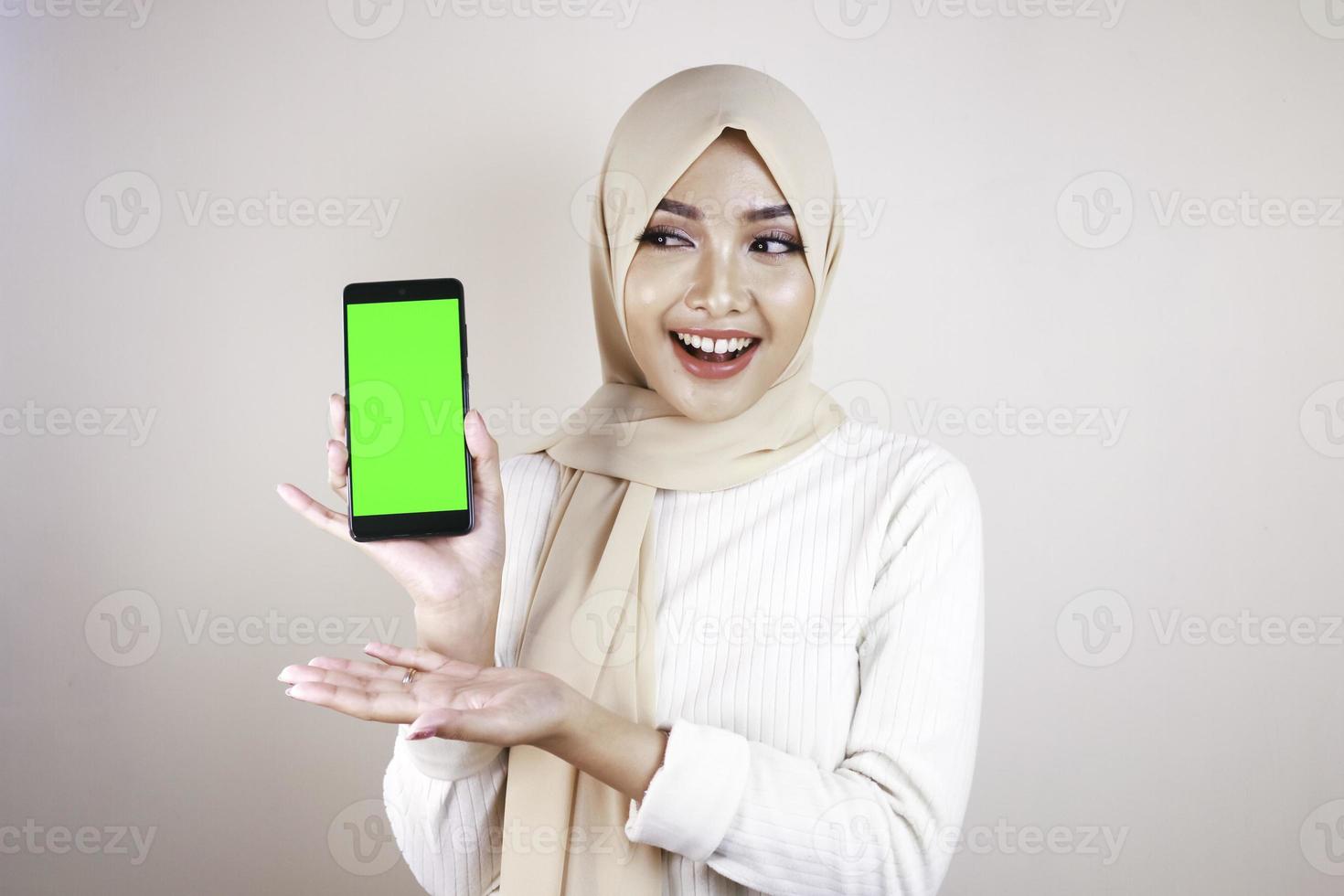 retrato de una alegre joven musulmana asiática apuntando a un celular con pantalla verde foto