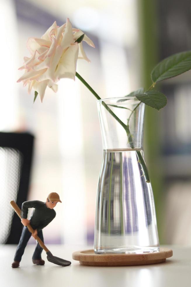 White rose in a glass vase with miniature worker doll holding shovel on table, miner man at work tiny figure toy model digging ground or gardening photo