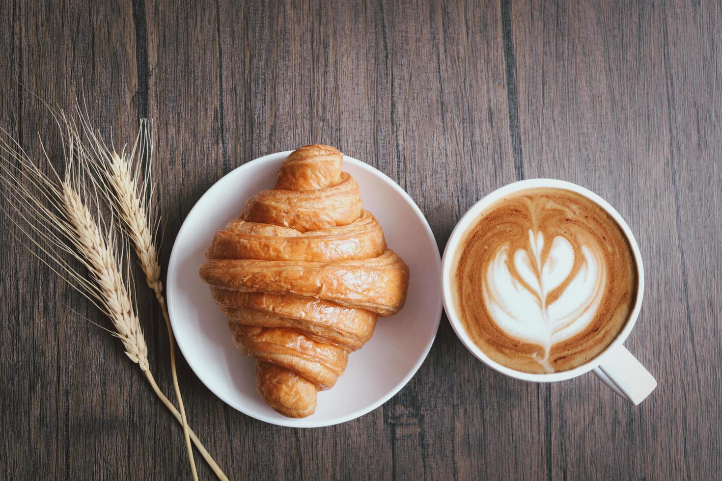 Freshly baked delicious croissant with spikelets and cup of beautiful morning coffee on wooden breakfast table, top view photo