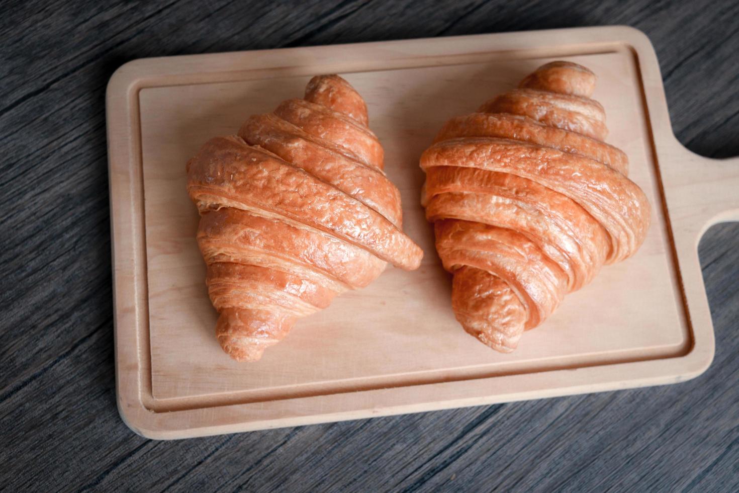 dos deliciosos cruasanes recién horneados en una tabla de cortar de madera, comida en la mesa del desayuno foto