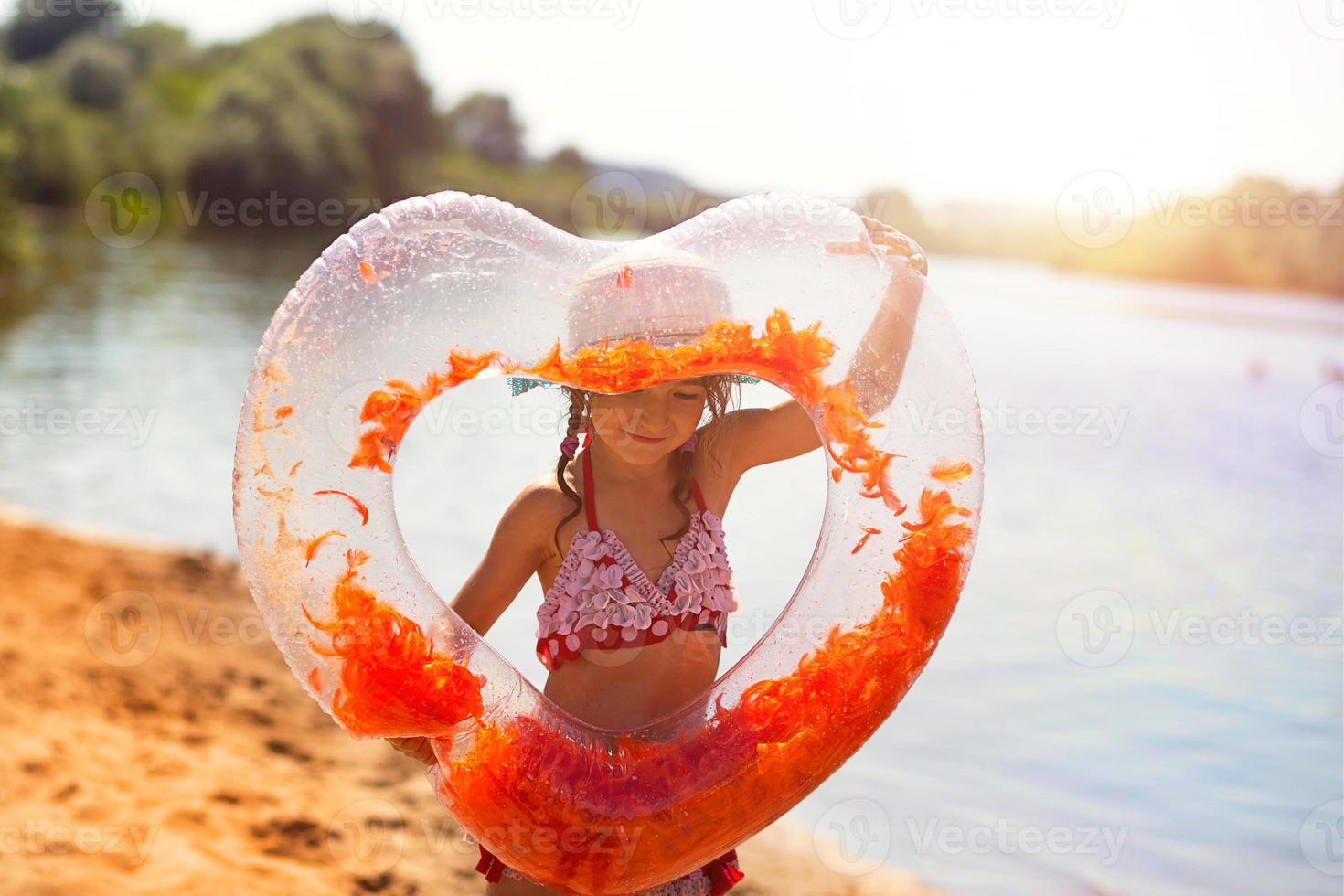 una chica con sombrero se encuentra en la orilla del río con un círculo inflable transparente en forma de corazón con plumas naranjas en el interior. vacaciones en la playa, nadar, broncearse, protectores solares. foto