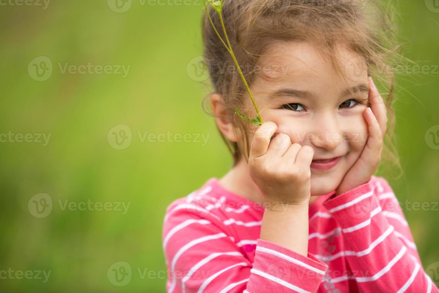 Little girl in a coral striped T-shirt on a green background in a field holds her face in her hands and smiles slyly. Children's Day, happy child, environmental and nature protection, insect repellent photo