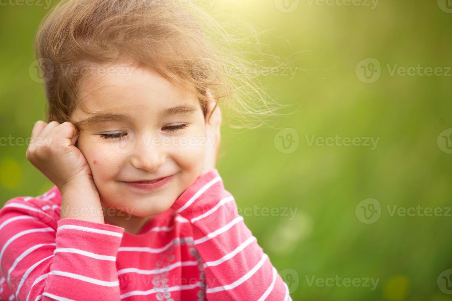 una niña pequeña con una camiseta a rayas de coral sobre un fondo verde en un campo sostiene su cara en sus manos y sonríe astutamente. día del niño, niño feliz, protección del medio ambiente y la naturaleza, repelente de insectos foto