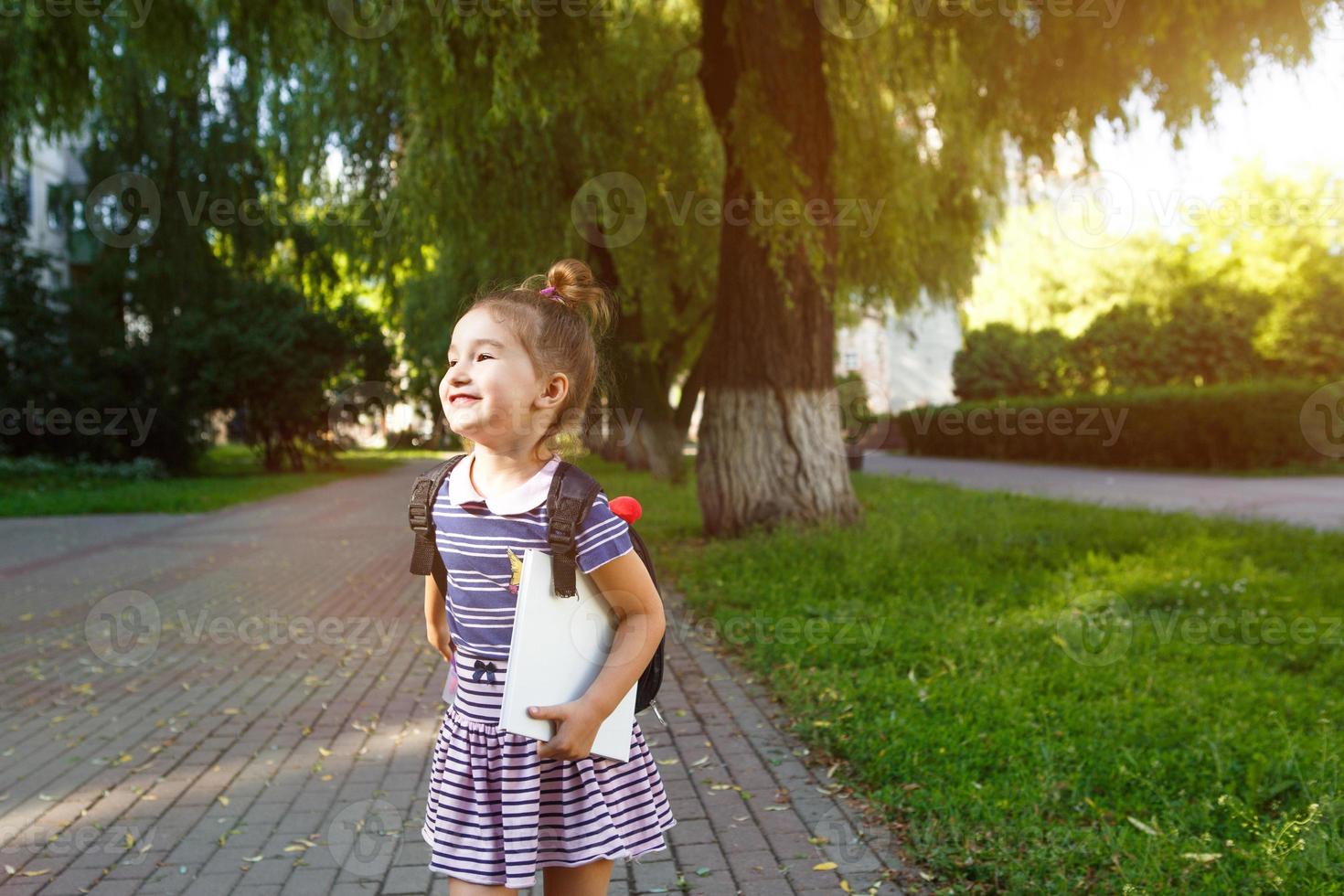 A little girl of Caucasian appearance in a school uniform with a backpack and the book. Concept back to school. Elementary school, developing activities for preschoolers. Space for text photo